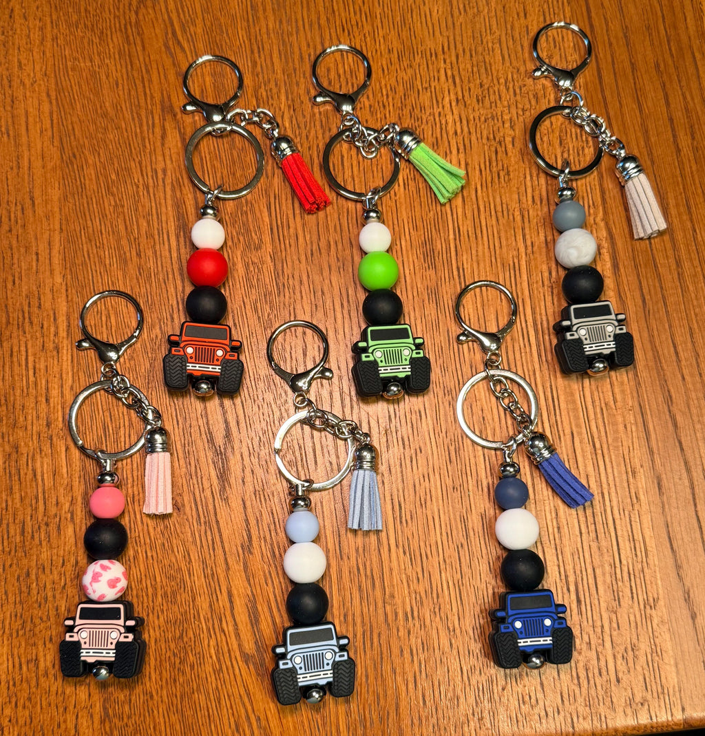 Six colorful beaded keychains with small vehicle charms are arranged on a wooden tabletop. Each keychain has a silver clasp, metal key ring, black and white beads, a colored bead, a tassel, and off-road vehicle charm in red, green, black, pink, blue, and white.