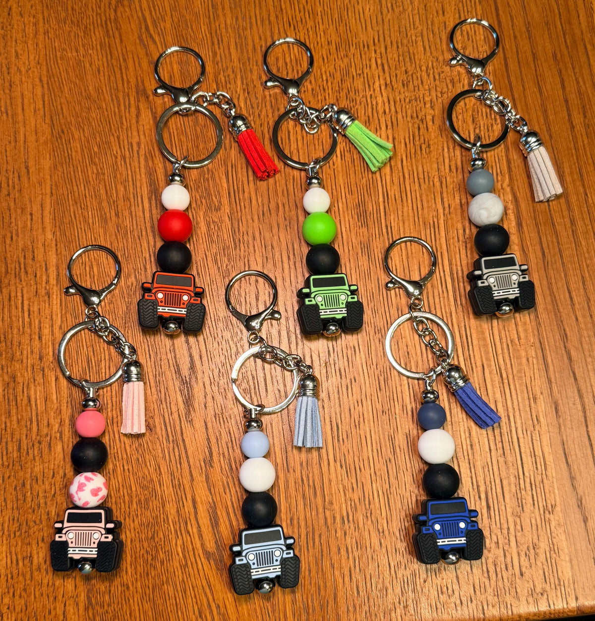 Six colorful beaded keychains with small vehicle charms are arranged on a wooden tabletop. Each keychain has a silver clasp, metal key ring, black and white beads, a colored bead, a tassel, and off-road vehicle charm in red, green, black, pink, blue, and white.