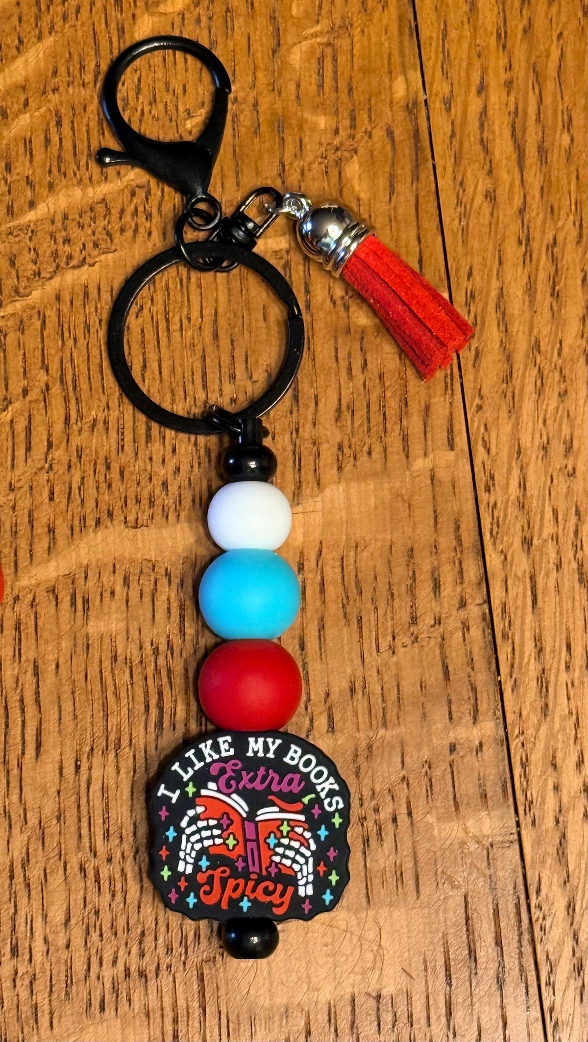 A beaded keychain with a black metal split ring, a red tassel, and four matte beads in white, light blue, red, and black. The bottom charm is a round black plaque featuring a colorful book illustration and the text 'I LIKE MY BOOKS Extra Spicy'.