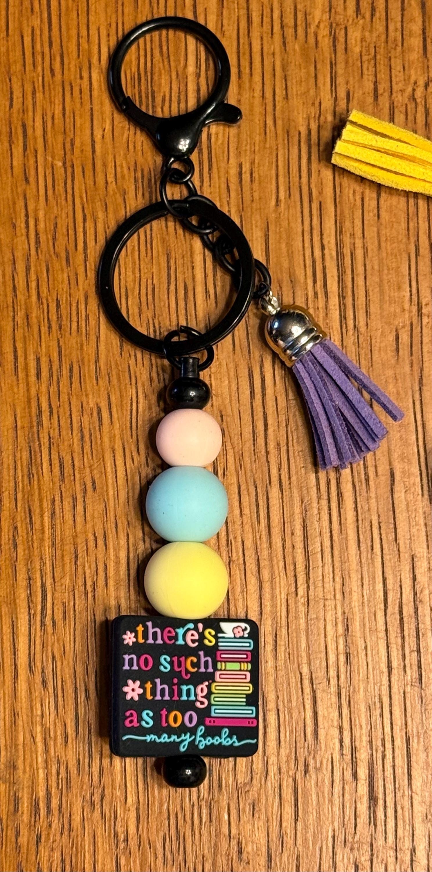 A black keychain with a split ring, a purple tassel, pastel-colored beads, and a small black charm that reads 'There's no such thing as to many books on a wooden background.