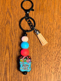 A black metal keychain with a clasp, a black chain, a light wood tassel, and a beaded charm featuring a blue book bag illustration and the text 'UH OH.. I bought more books