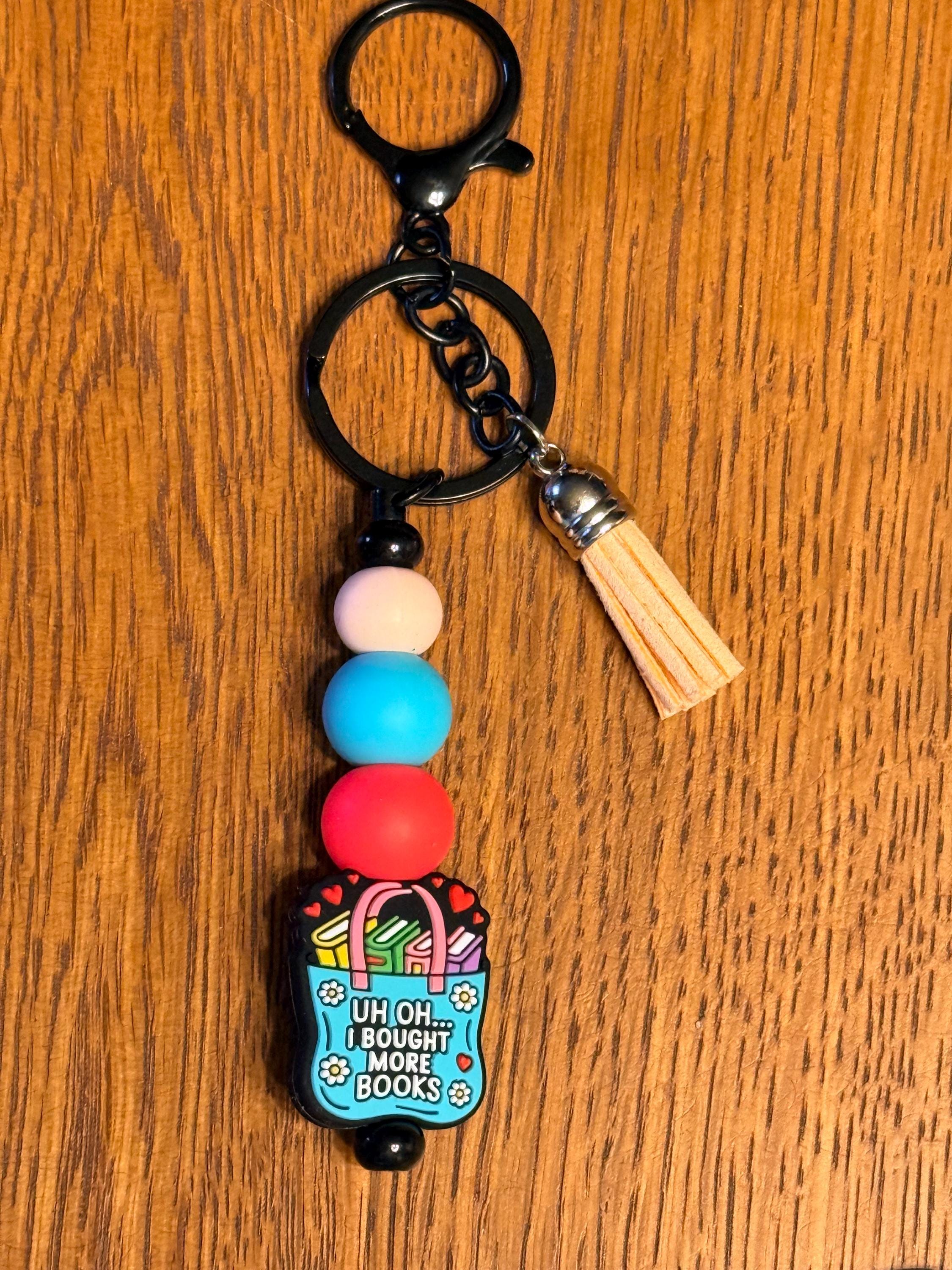 A black metal keychain with a clasp, a black chain, a light wood tassel, and a beaded charm featuring a blue book bag illustration and the text 'UH OH.. I bought more books
