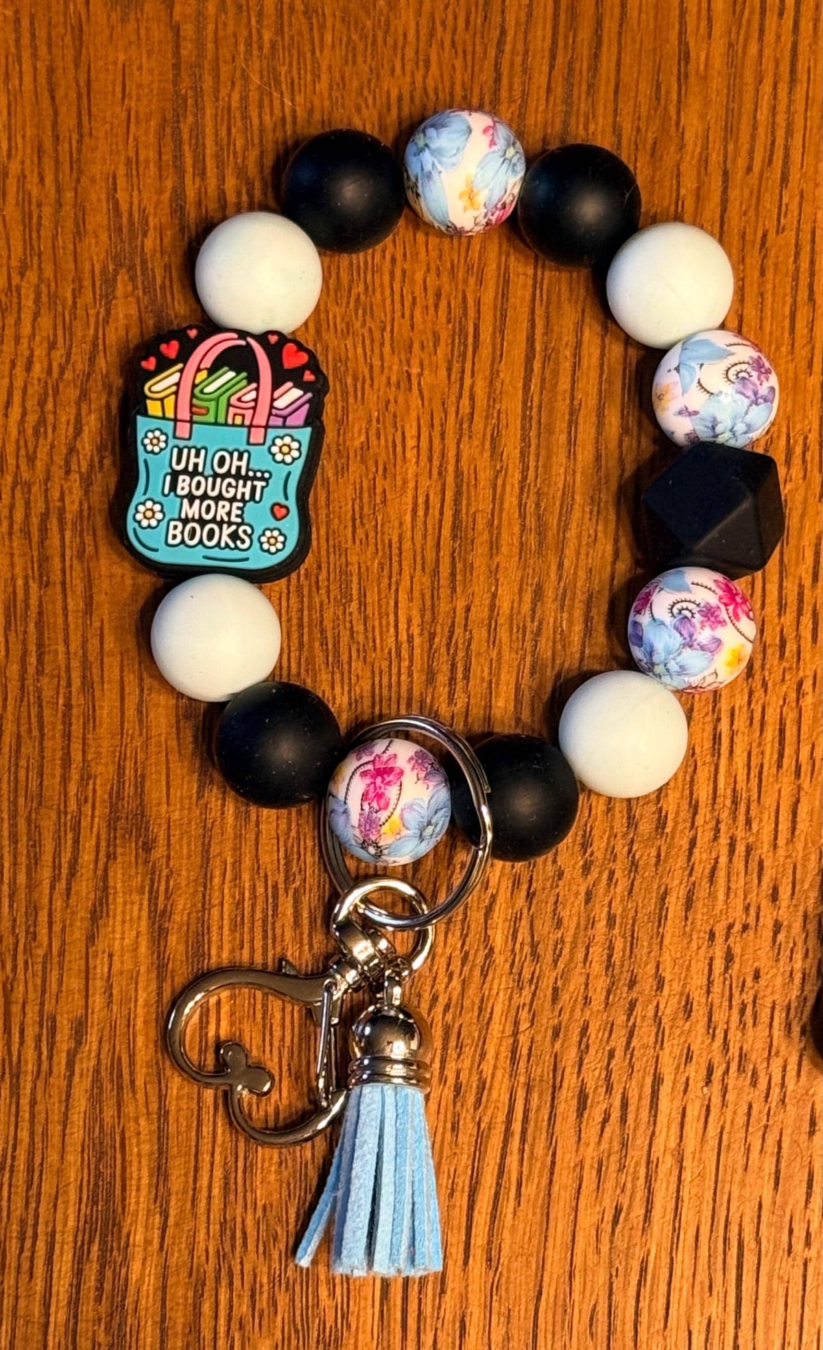 A beaded bracelet keychain on a wooden surface, featuring white and black beads, marbled beads, a small heart-shaped charm that reads 'UH OH...I bought more books.