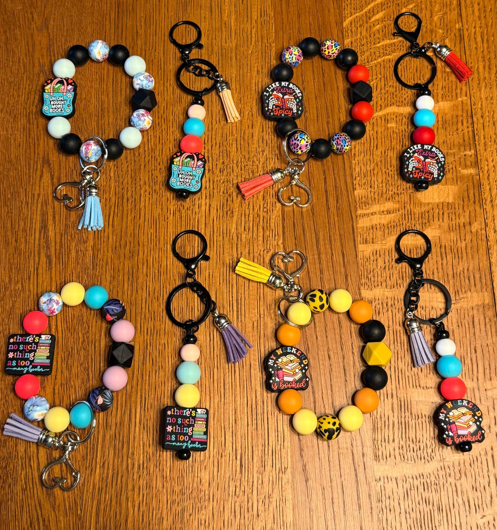 Six colorful beaded keychains with cartoon character charms are arranged on a wooden surface. Each keychain features black and multicolor beads, a decorative charm, and a tassel or clasp. Books, Reading theme