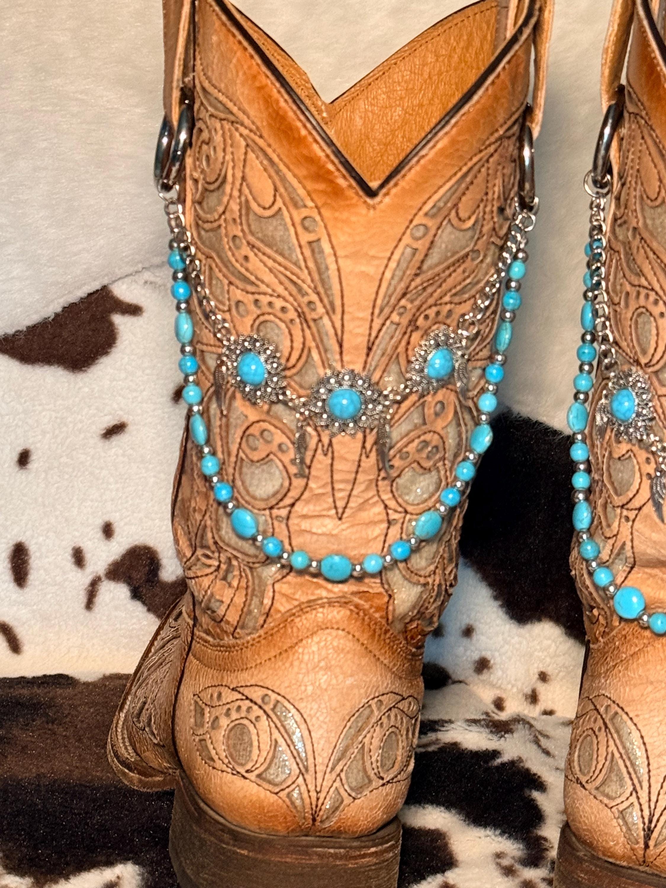 A close-up of a tan leather cowboy boot with intricate hand-tooled western patterns, on cowhide rug with turquoise bead and silver spacer bead boot bracelet chains and large silver filigree flower concho centerpiece with turquoise cabochon stones, Western boho boot jewelry