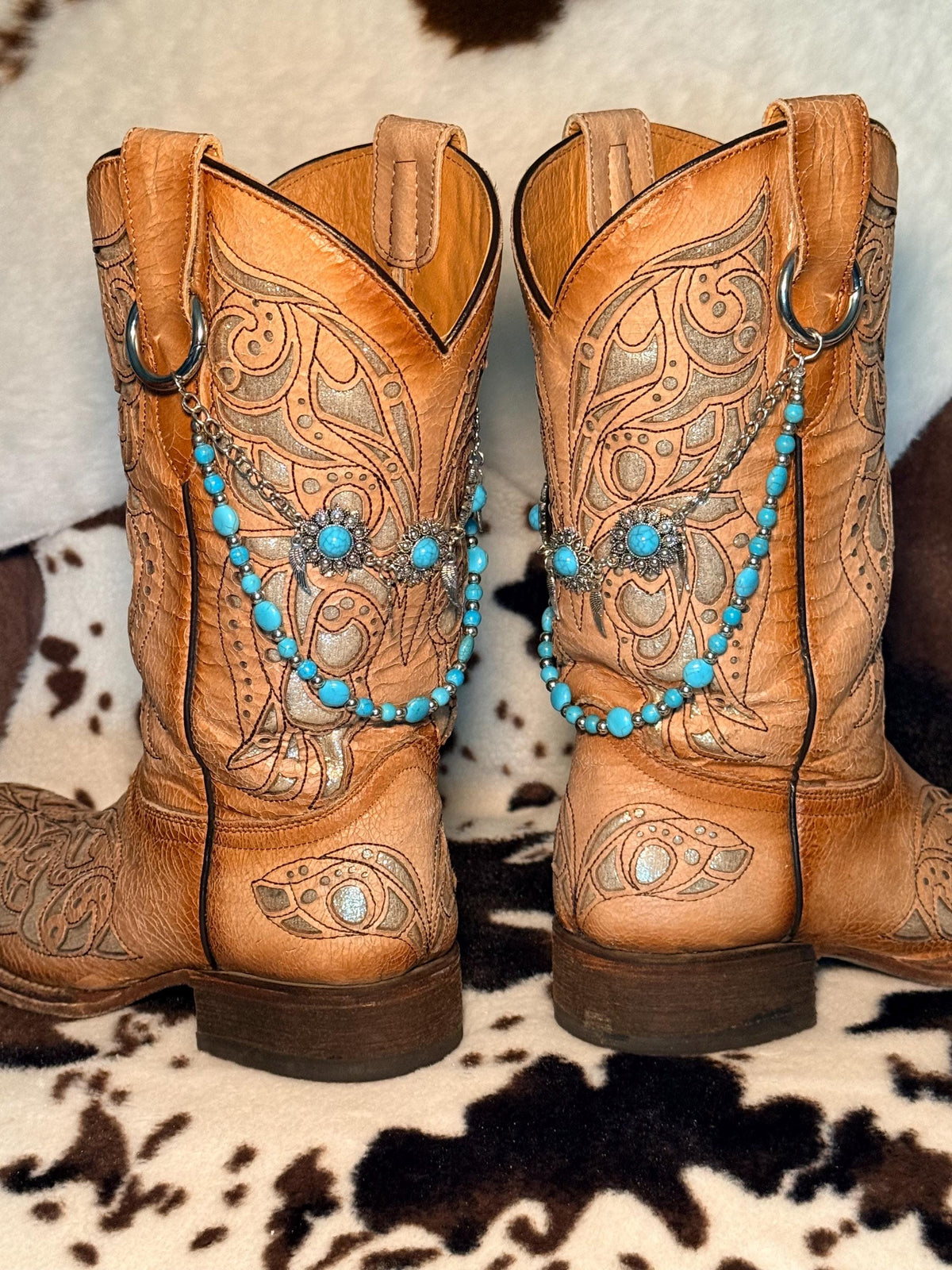 A pair of tan leather western cowboy boots with intricate floral tooling on cowhide rug with turquoise bead and silver spacer bead boot bracelet chains and large silver filigree flower concho centerpiece, Western boho boot bling set of 2