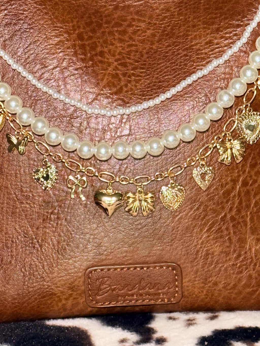 Close-up of brown distressed Bandana leather shoulder bag with three-strand Western boho purse chain showing faux seed pearl bead strand, larger faux round pearl bead strand, and gold oval link charm chain with nine gold charms including large puffy heart at center, large gold bow, ornate filigree hearts, crystal hearts, and small gold bows, Western boho handbag necklace detail