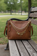 Front view of brown distressed Bandana leather shoulder bag on cowhide rug with three-strand Western boho purse chain featuring faux seed pearl bead strand, gold curb chain, and large gold oval link charm chain with eight heart and cross charms including white enamel cross, ornate gold cross, crystal rhinestone cross, gold hearts, and gold sunburst medallion