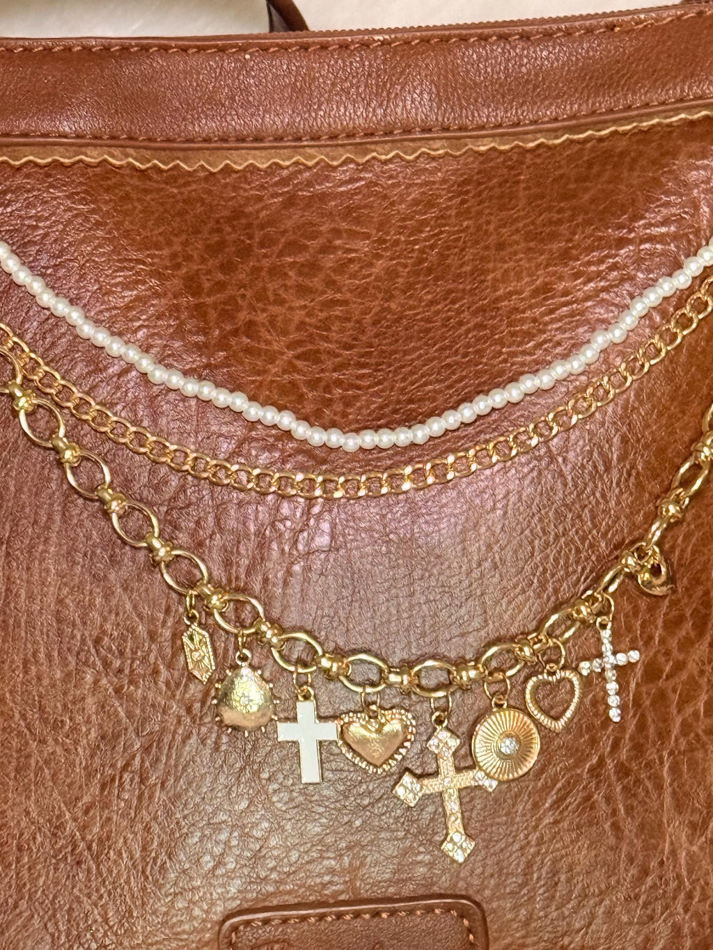Close-up of brown distressed Bandana leather shoulder bag with three-strand Western boho purse chain showing faux seed pearl bead strand, gold curb chain, and large gold oval link charm chain with eight charms: gold shield tag, gold teardrop heart, white enamel cross, gold ornate heart with rope border, large ornate gold cross, gold sunburst medallion, open gold heart, and crystal rhinestone cross