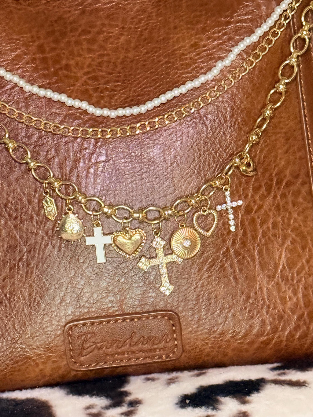 Alternate close-up of brown distressed Bandana leather shoulder bag on cowhide rug with three-strand Western boho purse chain featuring faux seed pearl bead strand, gold curb chain, and large gold oval link charm chain with eight heart and cross charms including crystal rhinestone cross, ornate gold cross, white enamel cross, gold ornate heart, and gold sunburst medallion, Western boho handbag accessory