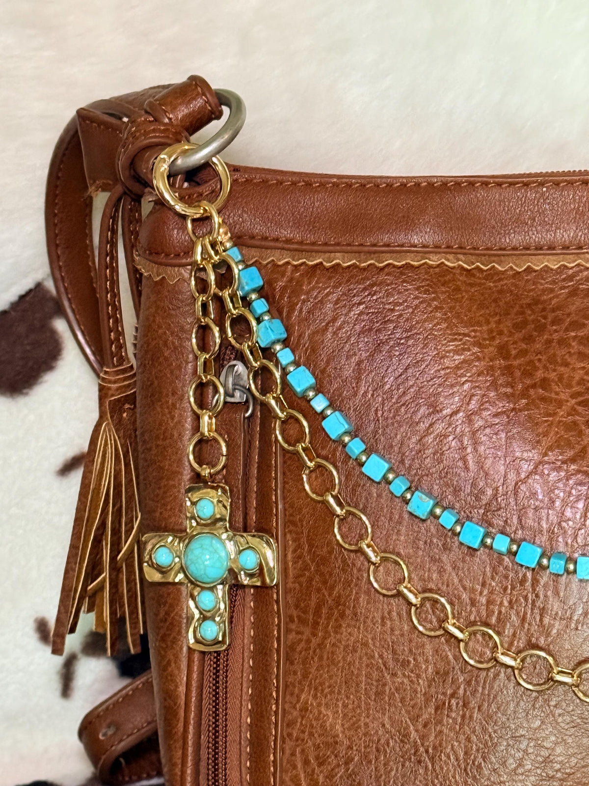 Close-up of left side hardware of brown distressed Bandana leather crossbody bag showing large ornate gold cross pendant with turquoise cabochon center stone and four corner stones hanging from gold ring clip, with turquoise square cube bead strand with gold rondelle spacers and bold gold round link chain across bag front, Western boho purse chain detail