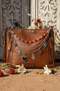 Brown distressed Bandana leather crossbody bag on sandy ground against ornate white carved wood backdrop with dried wildflowers, featuring three-strand Western boho purse chain with mixed red faceted oval bead and clear crystal bead strand, delicate silver cable chain, and silver curb chain with seven ornate silver sacred heart charms including black enamel and red enamel hearts, Western boho handbag accessory