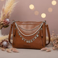 Front view of brown distressed Bandana leather shoulder bag on cowhide rug with three-strand silver Western boho purse chain featuring faux seed pearl bead strand, silver curb chain, and silver oval link charm chain with nine silver charms including bows, butterflies, ornate hearts, and crystal hearts, Western boho handbag accessory