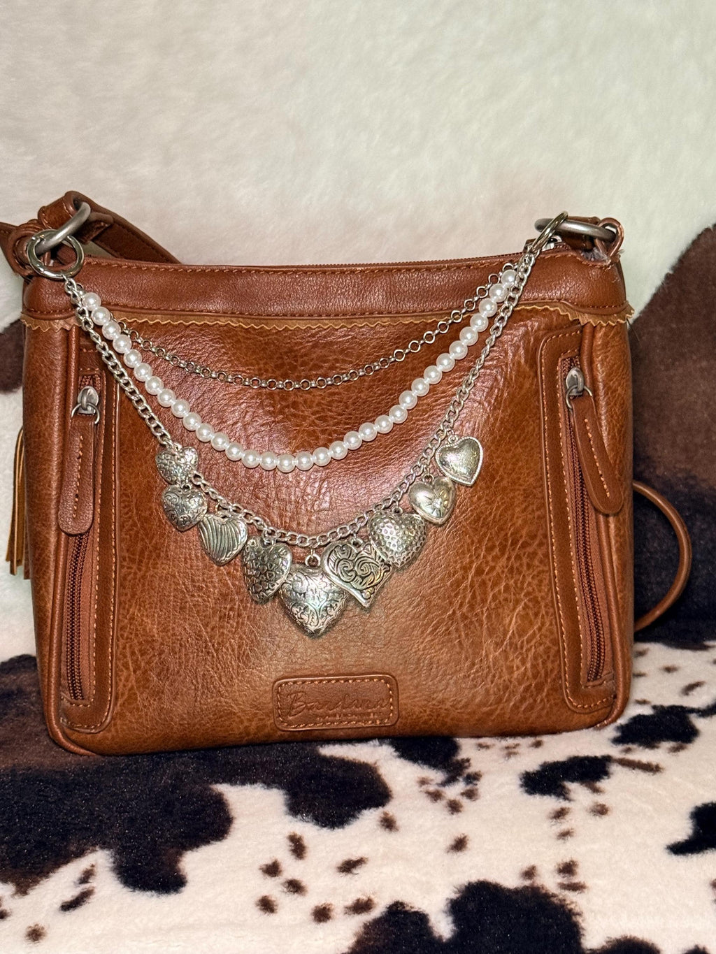 Alternate front view of brown distressed Bandana leather crossbody bag on cowhide rug with three-strand Western boho purse chain featuring delicate silver cable chain, faux uniform round pearl bead strand, and silver curb chain with eight large ornate silver heart charms including ribbed heart, floral scroll engraved heart, large filigree heart at center, Celtic swirl heart, and hammered texture heart