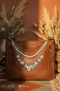 Front view of brown distressed Bandana leather crossbody bag with three-strand Western boho purse chain featuring delicate silver cable chain, faux uniform round pearl bead strand, and silver curb chain with eight large ornate silver heart charms in varying engraved and textured styles including large filigree heart at center, Western boho handbag accessory