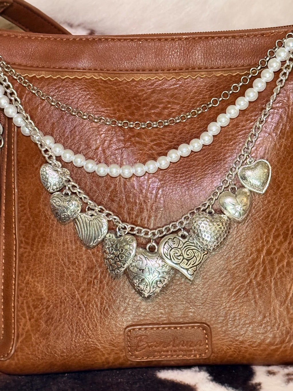 Close-up of brown distressed Bandana leather crossbody bag with three-strand Western boho purse chain showing delicate silver cable chain, faux uniform round pearl bead strand, and silver curb chain with eight large ornate silver heart charms including large filigree engraved heart at center, Celtic swirl engraved heart, hammered texture heart, ribbed heart, floral scroll heart, puffed star-center heart, and smooth locket-style heart, Western boho handbag necklace detail