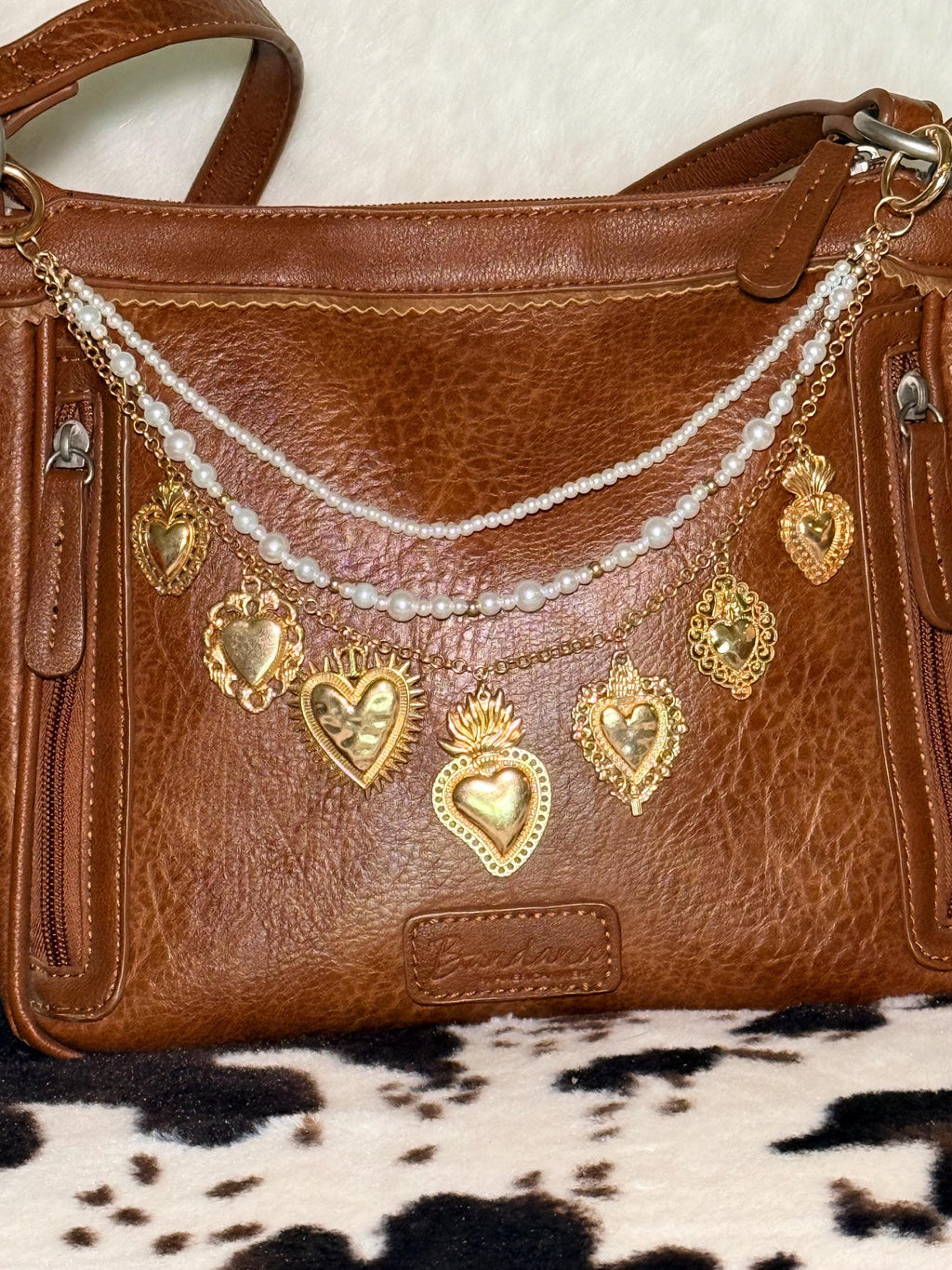 Front view of brown distressed Bandana leather shoulder bag on cowhide rug with three-strand Western boho purse chain featuring faux seed pearl bead strand, delicate gold cable chain, and gold curb chain with seven large ornate gold sacred heart charms including sunburst radiating heart, flaming sacred heart, filigree-bordered heart, crown heart, and milagro-style hearts