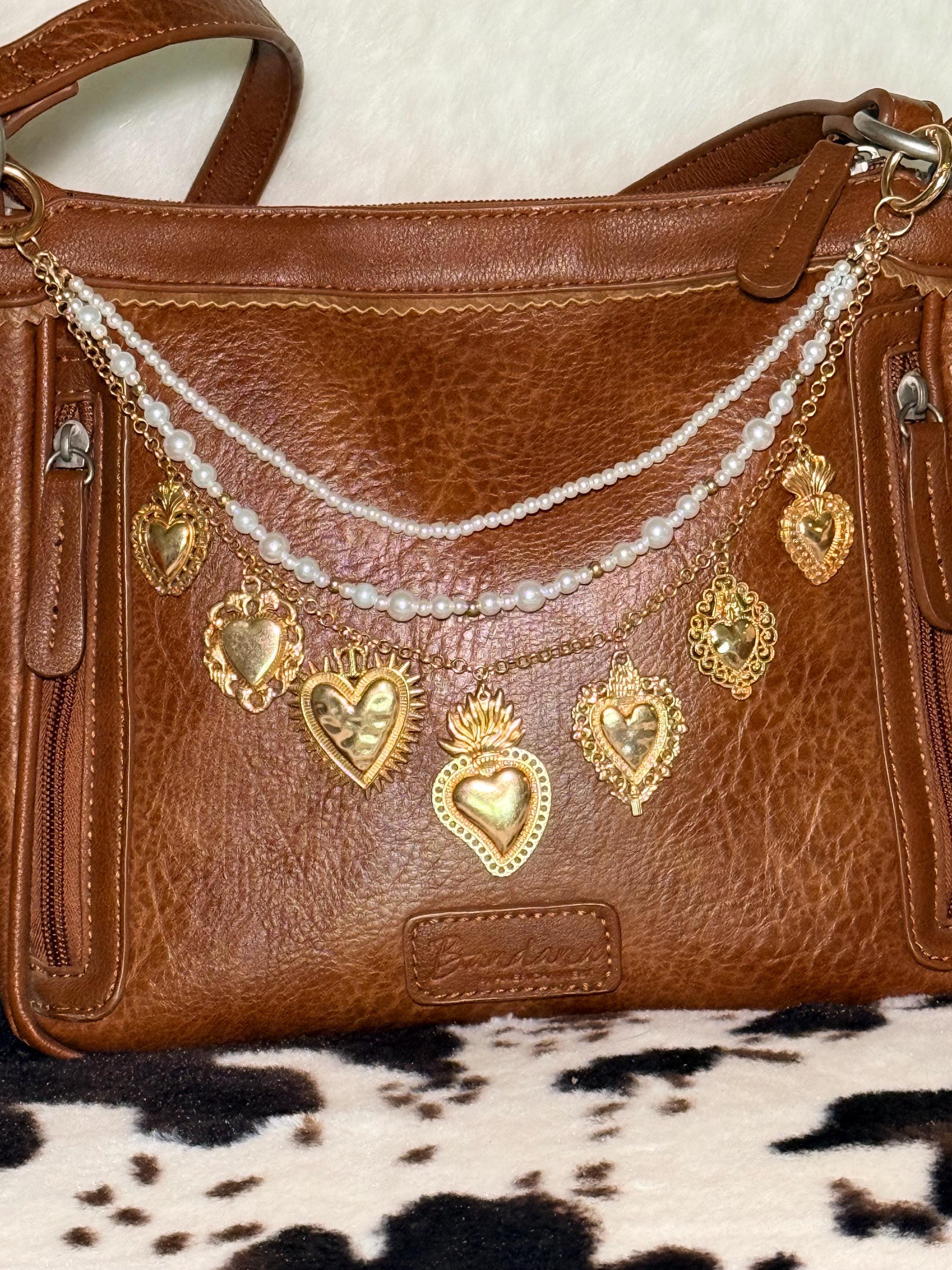 Front view of brown distressed Bandana leather shoulder bag on cowhide rug with three-strand Western boho purse chain featuring faux seed pearl bead strand, delicate gold cable chain, and gold curb chain with seven large ornate gold sacred heart charms including sunburst radiating heart, flaming sacred heart, filigree-bordered heart, crown heart, and milagro-style hearts