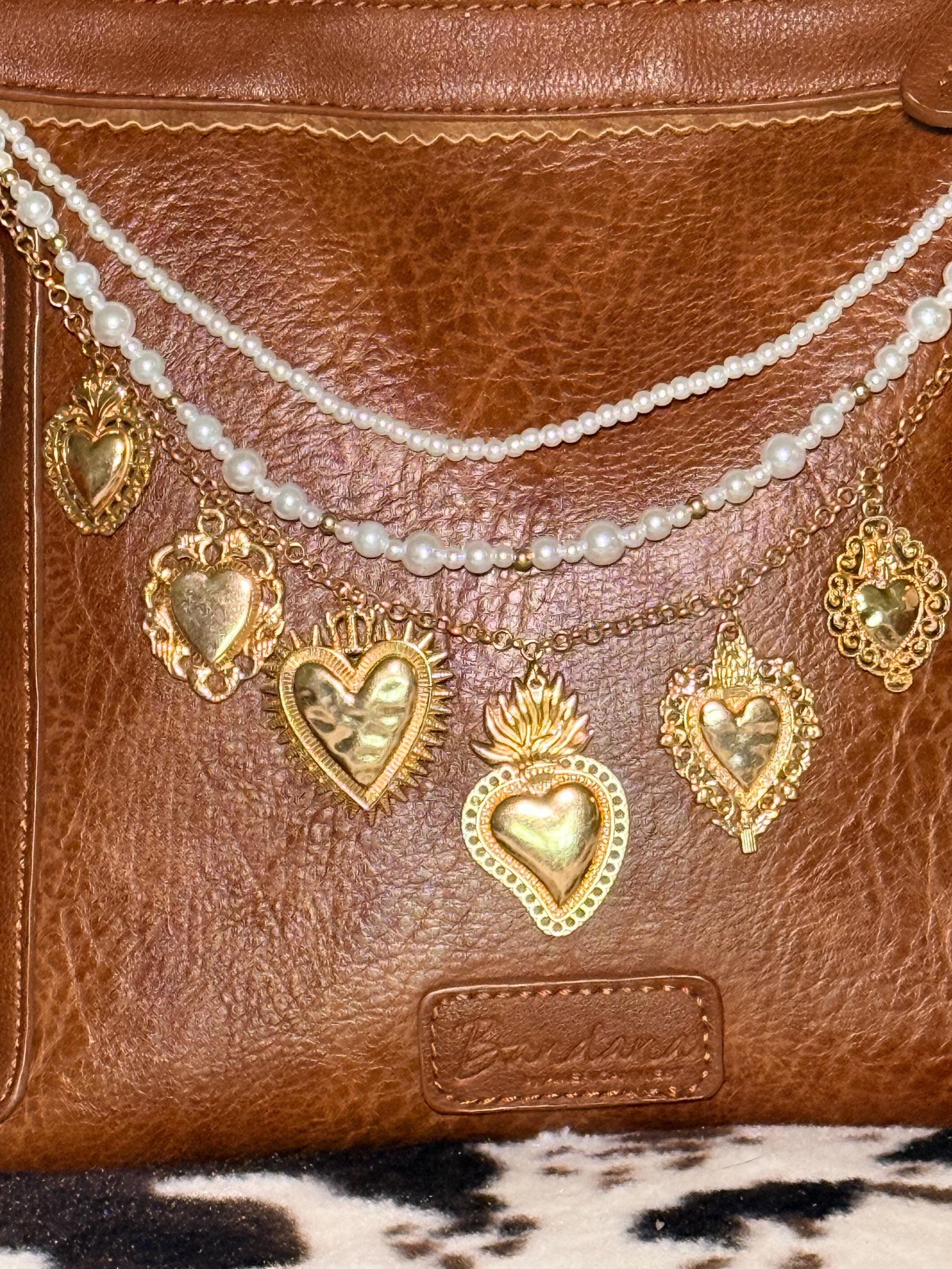Close-up of brown distressed Bandana leather shoulder bag with three-strand Western boho purse chain showing faux seed pearl bead strand, delicate gold cable chain, and gold curb chain with seven large ornate gold sacred heart charms including sunburst radiating heart, flaming sacred heart, filigree-bordered heart, crown heart, and ornate milagro-style hearts, Western boho handbag necklace detail