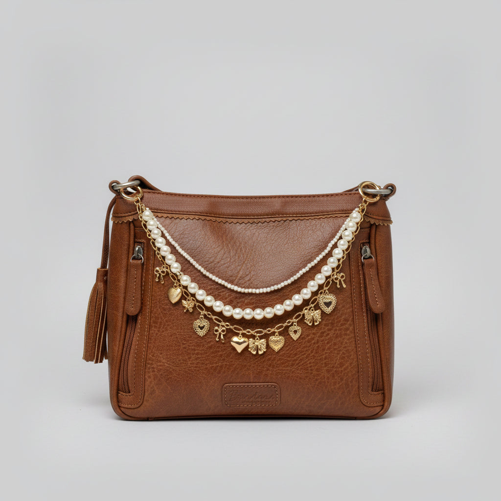 Front view of brown distressed Bandana leather shoulder bag on cowhide rug with three-strand Western boho purse chain featuring faux seed pearl bead strand, larger faux round pearl bead strand, and gold oval link charm chain with nine gold heart and bow charms including large puffy heart and large bow at center, Western boho handbag accessory