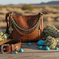 Front view of brown distressed Bandana leather crossbody bag with two-strand Western boho purse chain featuring turquoise square cube bead strand with gold rondelle spacers above and bold gold round link chain below, with large ornate gold cross pendant with turquoise cabochon stones hanging from left side hardware, Western boho handbag accessory