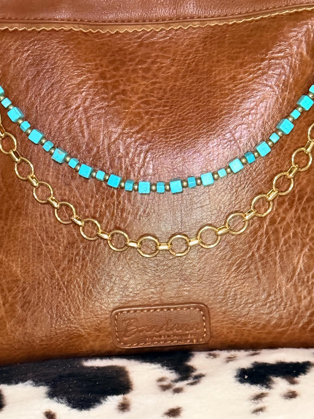 Close-up of brown distressed Bandana leather crossbody bag showing two-strand Western boho purse chain with turquoise square cube bead strand with small gold rondelle spacers between each bead above and bold gold round link chain below, layered Western boho handbag accessory detail
