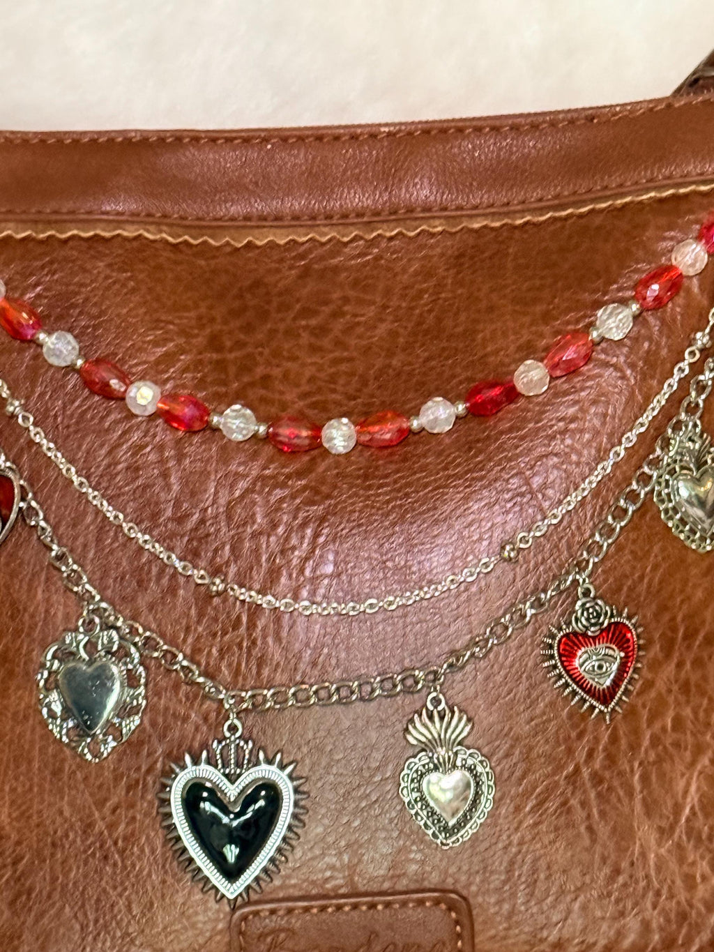 Close-up of brown distressed Bandana leather crossbody bag with three-strand Western boho purse chain showing mixed red faceted oval bead and clear crystal bead strand, delicate silver cable chain, and silver curb chain with seven silver sacred heart charms including black enamel sunburst heart with crown, silver flaming sacred heart, red enamel sacred heart with eye detail and rose, and silver mirror-finish ornate heart, Western boho handbag necklace detail