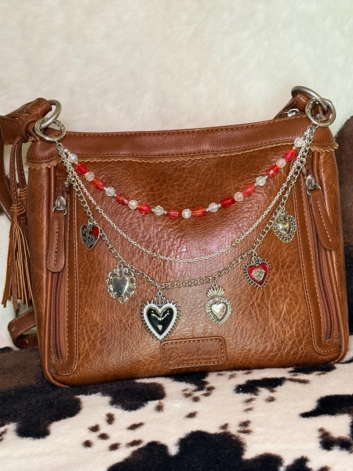 Front view of brown distressed Bandana leather crossbody bag on cowhide rug with three-strand Western boho purse chain featuring mixed red faceted oval bead and clear crystal bead strand, delicate silver cable chain, and silver curb chain with seven ornate silver sacred heart charms including black enamel sunburst heart with crown, red enamel sacred heart with eye detail and rose, filigree-bordered silver heart, and silver flaming sacred heart