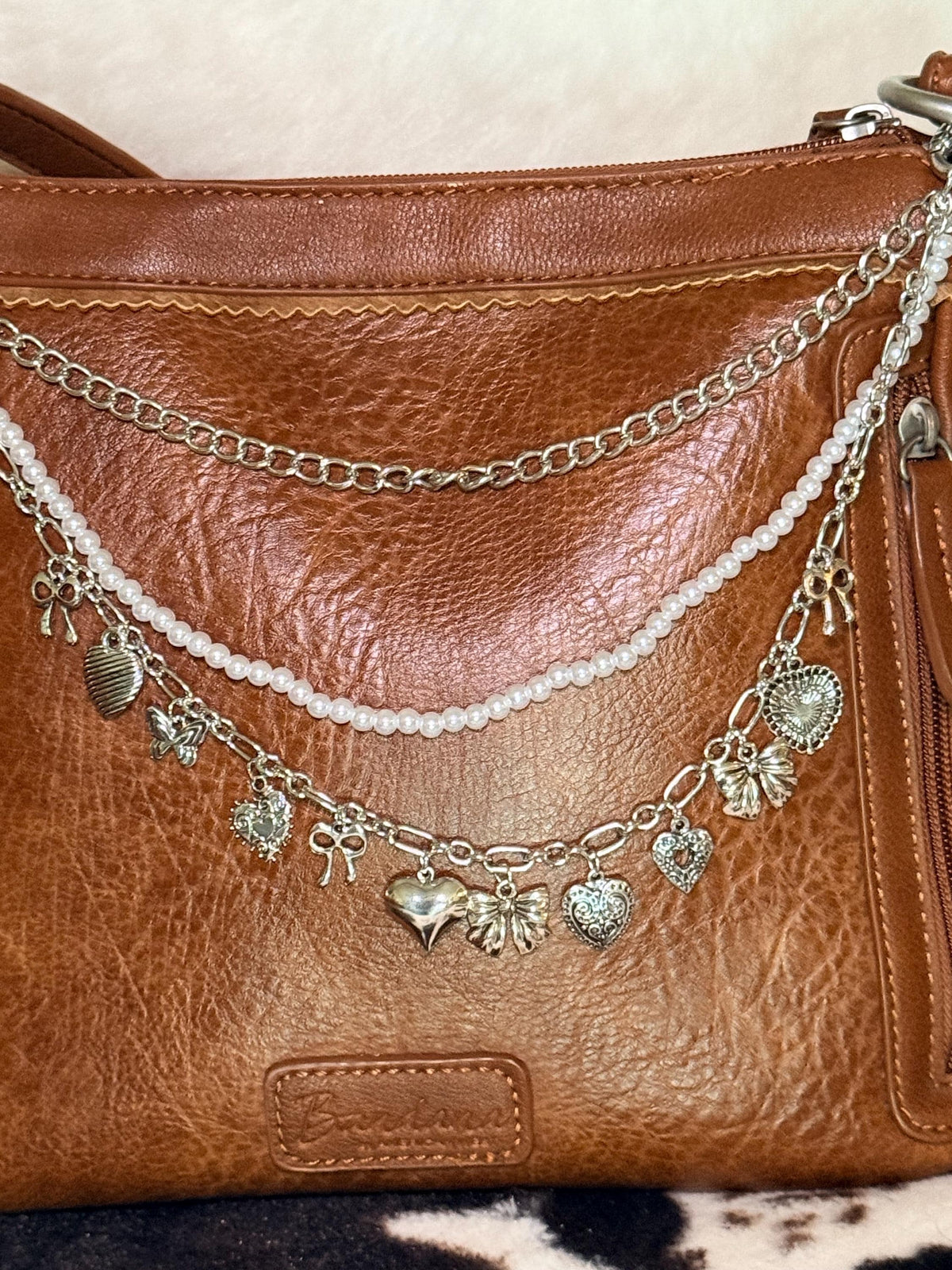 Close-up of brown distressed Bandana leather shoulder bag with three-strand silver Western boho purse chain showing faux seed pearl bead strand, silver curb chain, and silver oval link charm chain with nine silver charms including large silver butterfly bow at center, ornate filigree hearts, crystal hearts, and silver tag charm, Western boho handbag necklace detail