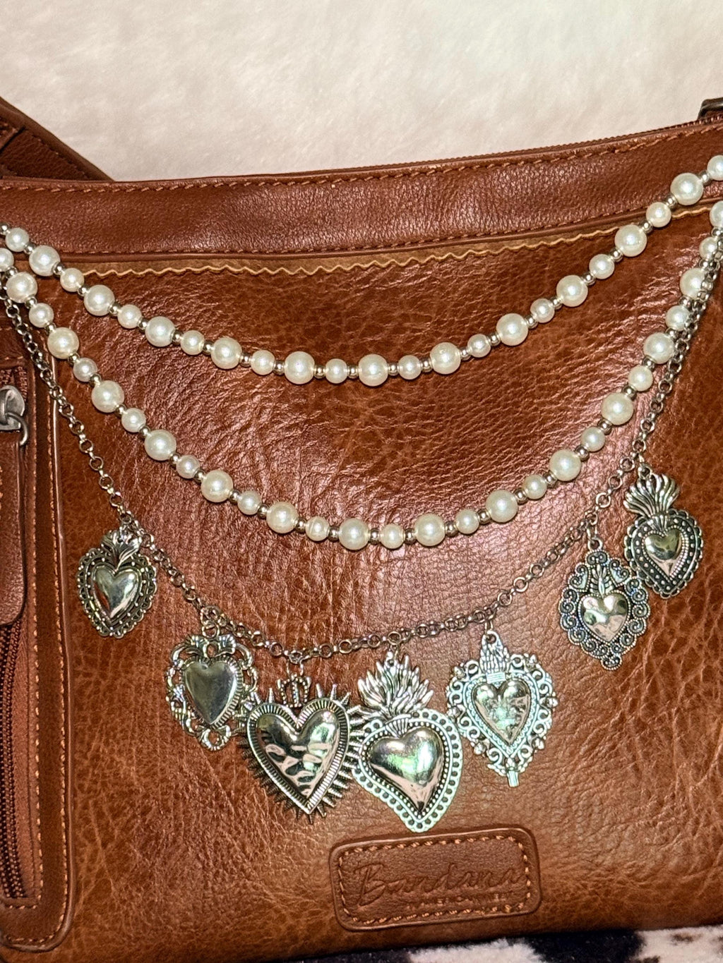 Close-up of brown distressed Bandana leather crossbody bag with four-strand Western boho purse chain showing small and larger faux pearl bead strands, delicate silver cable chain, and silver curb chain with seven large ornate silver sacred heart charms: large flaming sacred heart with sunburst rays at center, large flaming sacred heart with wings, filigree-bordered sacred heart with flower detail, filigree scroll heart, smooth puffed heart, small smooth heart, and flaming sacred heart with crown