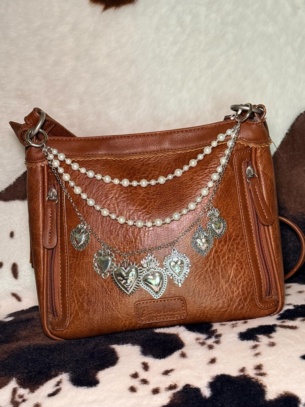 Alternate front view of brown distressed Bandana leather crossbody bag on cowhide rug with four-strand Western boho purse chain featuring small and larger faux pearl bead strands, delicate silver cable chain, and silver curb chain with seven large ornate silver sacred heart charms including large flaming sacred heart with sunburst rays at center and flaming sacred heart with wings, Western boho handbag accessory
