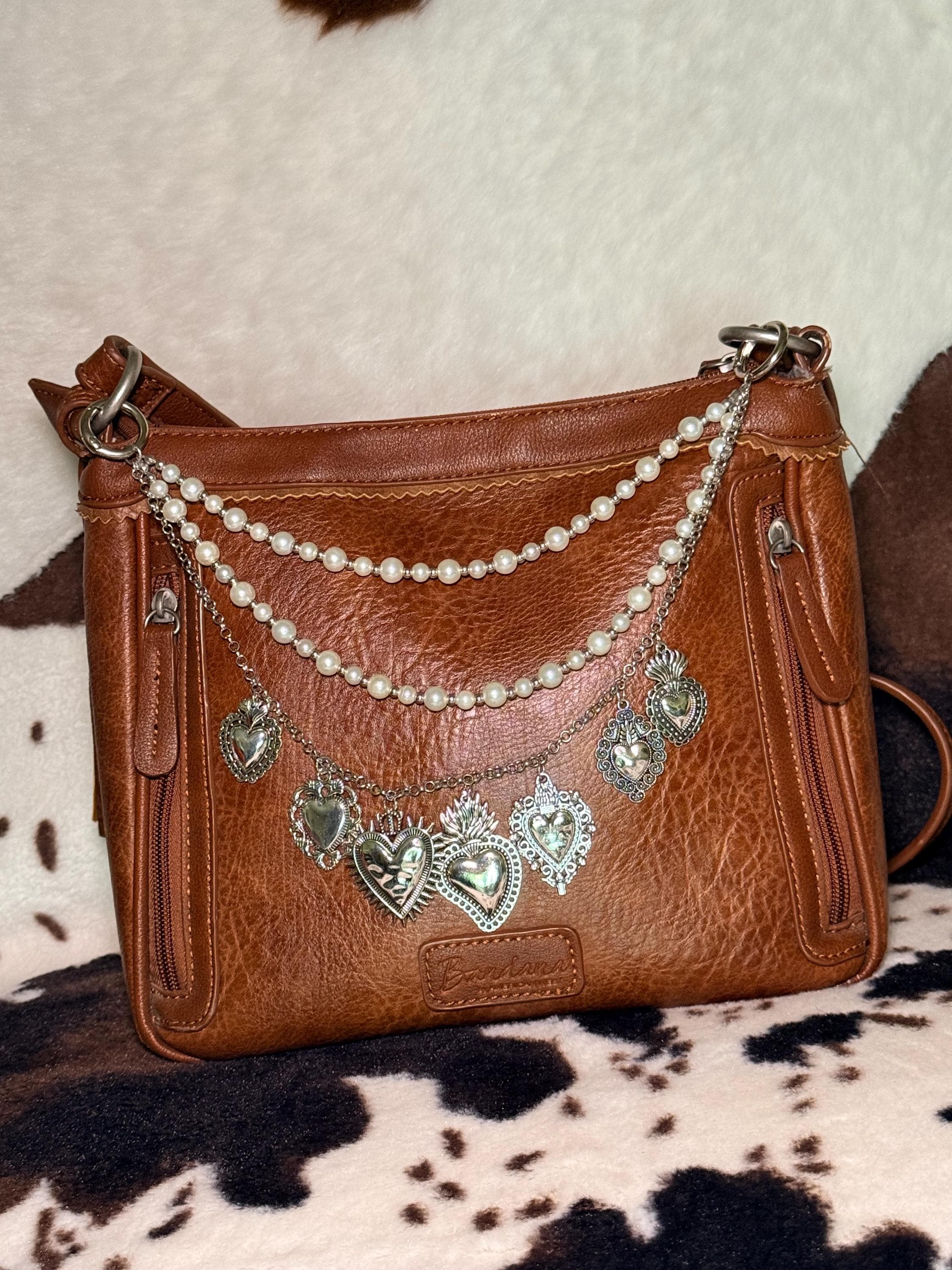 Alternate front view of brown distressed Bandana leather crossbody bag on cowhide rug with four-strand Western boho purse chain featuring small and larger faux pearl bead strands, delicate silver cable chain, and silver curb chain with seven large ornate silver sacred heart charms including large flaming sacred heart with sunburst rays at center and flaming sacred heart with wings, Western boho handbag accessory