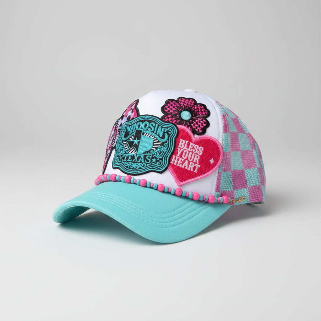 A colorful trucker-style cap with a white front panel, pink and light blue checkered mesh side panels, a turquoise brim, and decorative patches including a flower, a heart that says 'BLISS YOUR HEART,' and a turquoise badge. 