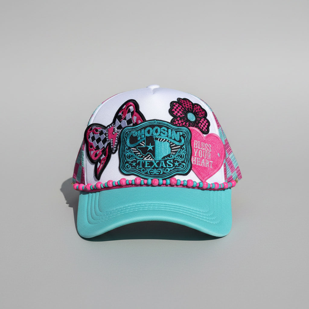 A turquoise and white trucker hat with decorative embroidered patches, including a checkered bow, flower, and a central Texas-themed patch.