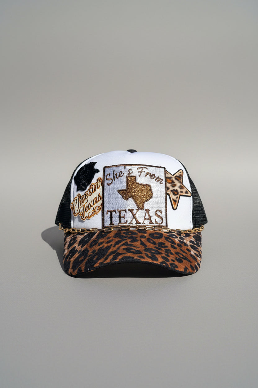 A trucker-style cap with a white front panel, black mesh sides, and a leopard-print brim. The front patch reads TEXAS' with a glittery Texas shape and a star motif, and the brim also shows a leopard print pattern.