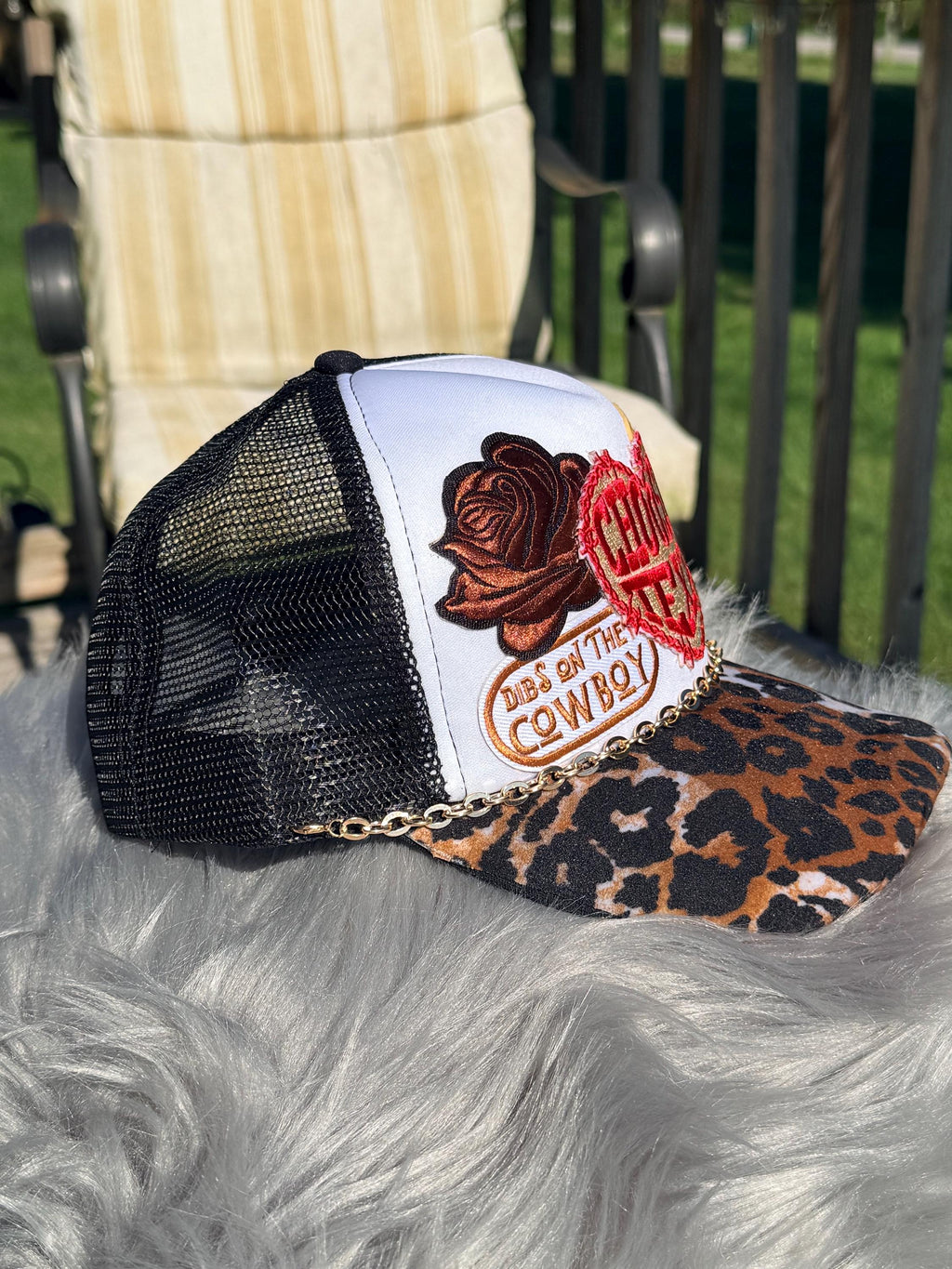 A trucker hat with a white front panel, black mesh back, leopard-print brim, and a decorative patch featuring a rose and the words 'COWBOY'. 