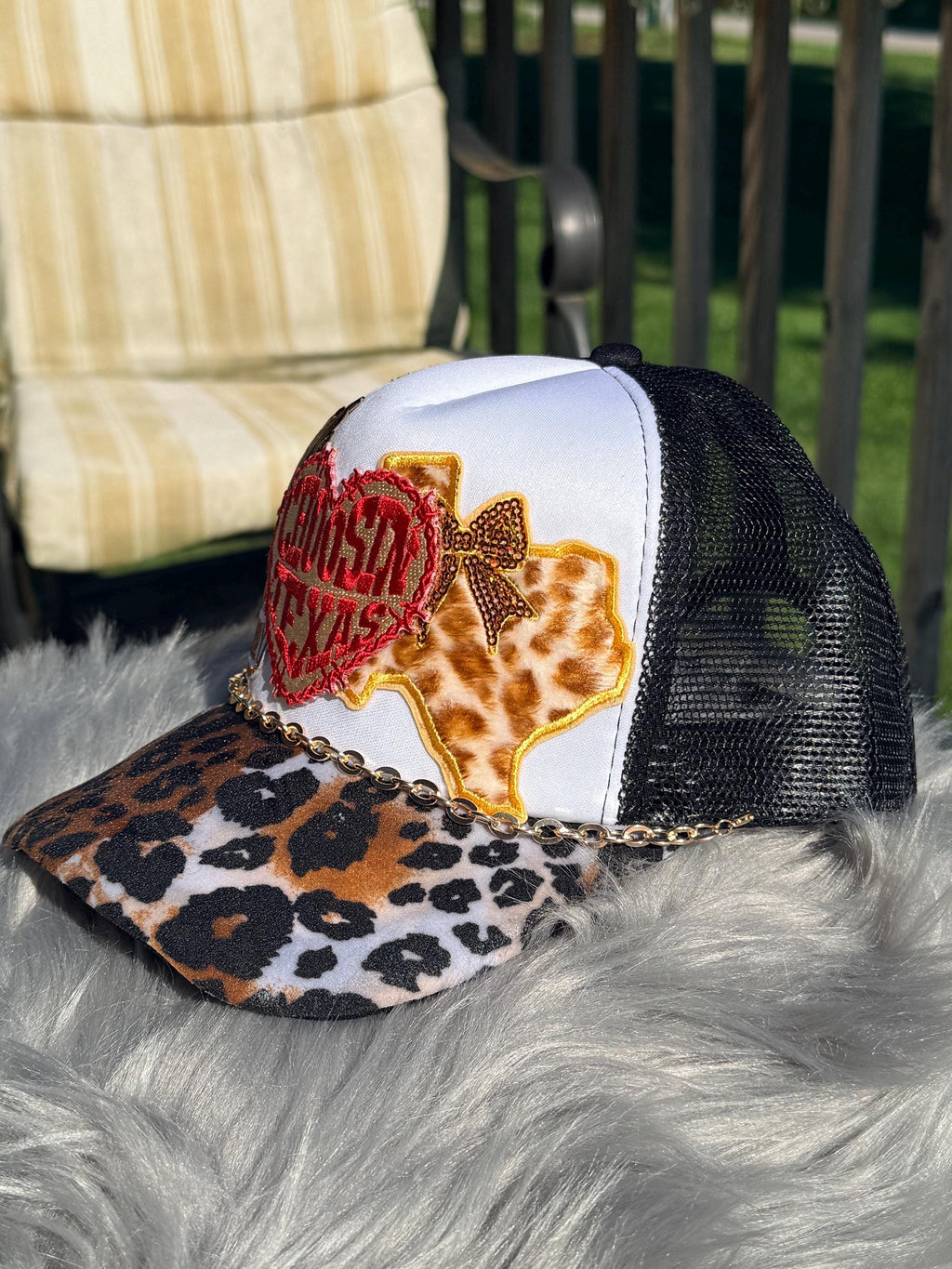A trucker-style cap with a white front panel, black mesh back, leopard-print brim, and decorative embroidered patches including a red heart and a Texas-shaped patch.