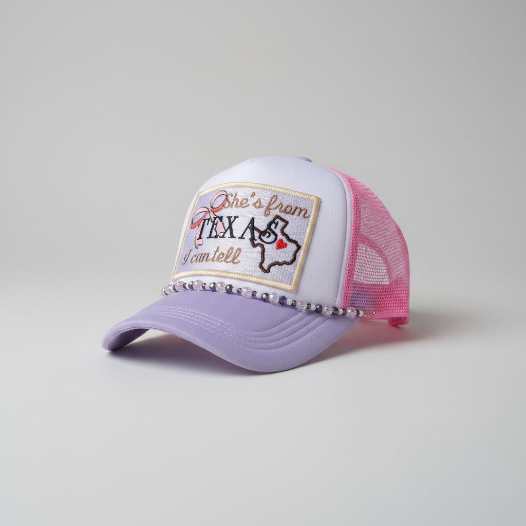 A lavender and pink trucker hat with a custom embroidered patch reading  Texas' and a beaded hat chain along the brim, 