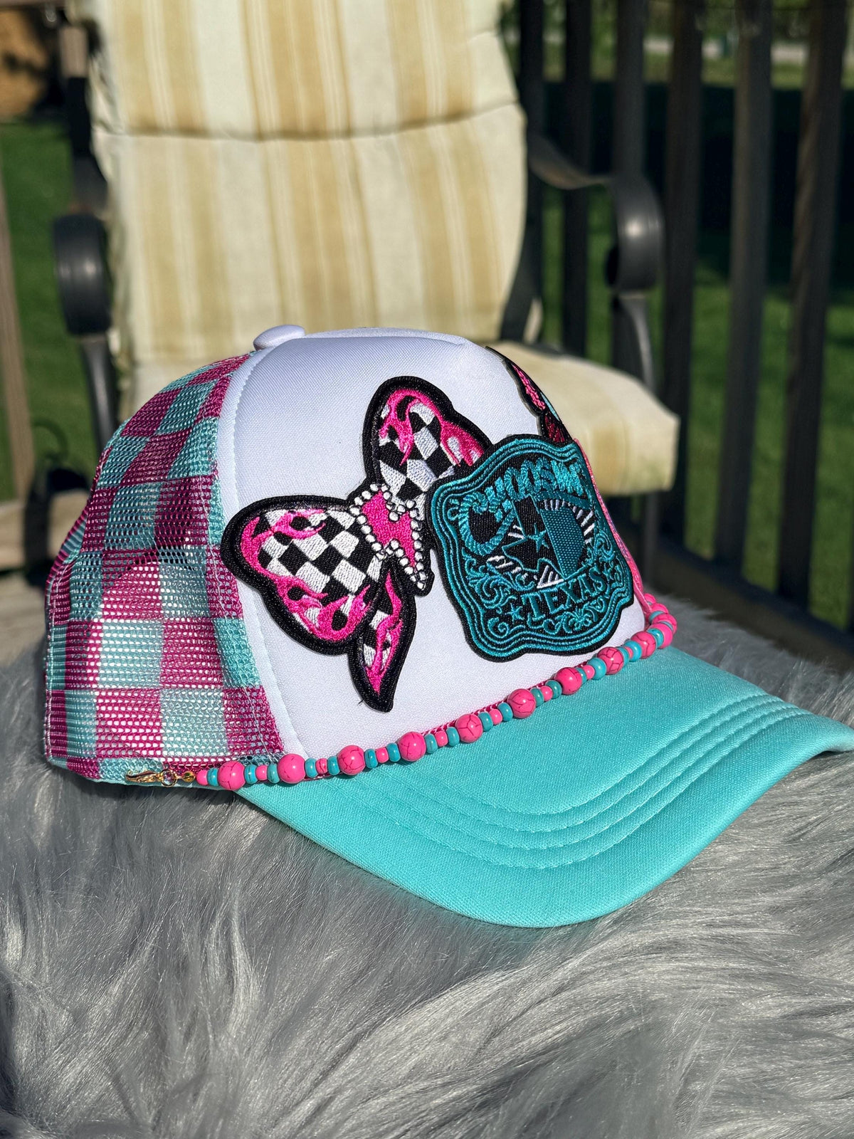A trucker-style cap with a white front panel, pink and teal plaid side panels, a pink-and-black checkered bow patch, a teal embroidered crest patch, and a turquoise brim with pink & turquoise hat chain.