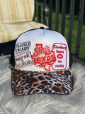 A trucker-style cap with a white front panel, black mesh sides, and a leopard-print visor,  The front has multiple embroidered-style patches including a large red Texas outline smaller text such as 'Trouble Maker,' 'Whiskey"  and 'Cowboy tears.